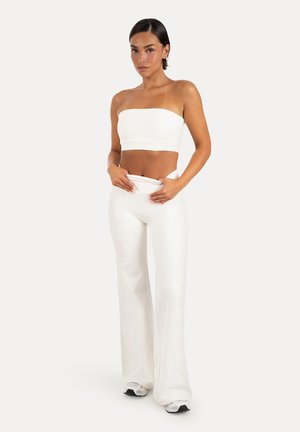 Kvinde iført en hvid stropløs crop top og højtaljede hvide bukser med vide ben, justerer linningen, stående på en ensfarvet baggrund.