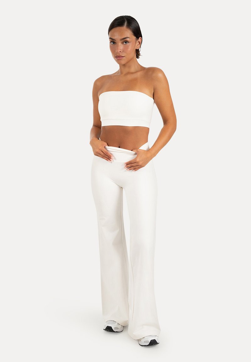 Femme portant un crop top blanc sans bretelles et un pantalon blanc taille haute à jambes larges, ajustant la taille, debout sur un fond uni.