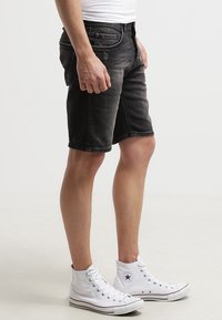 Persona vestida con pantalones cortos de mezclilla negros, zapatillas altas blancas y una camiseta blanca, de pie contra un fondo blanco.