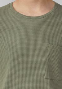 Oliwkowa koszulka z krótkim rękawem z teksturowanego materiału, z okrągłym dekoltem i kieszonką na piersi. Delikatny prążkowany wzór na całej powierzchni.