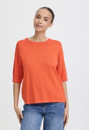 Femme à la peau brun clair et aux cheveux foncés, vêtue d'un haut orange à manches mi-longues et d'un jean bleu, debout devant un fond clair uni.