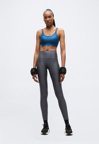 Αθλητική γυναίκα που στέκεται με εφαρμοστό μπλε αθλητικό σουτιέν, γκρι κολάν, μαύρα αθλητικά παπούτσια και βαράκια στα χέρια, μπροστά σε ένα απλό λευκό φόντο.