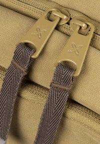Detailní záběr na hnědou látkovou tašku se dvěma béžovými zipy s logem a hnědými popruhy, zdůrazňující texturu a funkční design.