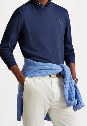 Hombre con camisa de manga larga azul marino, pantalones blancos, bufanda de lunares azul marino, cinturón marrón, reloj y suéter azul claro atado a la cintura.