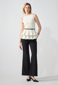 Kremasti peplum top s slojevitim volanima, uparen s crnim širim hlačama i crnim potpeticama. Sadrži uski crni remen i sjajnu narukvicu.