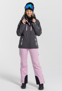 Grijze waterdichte jas met roze ritsen, gecombineerd met lichtroze broeken met zwarte accenten. Model met een zwarte beanie en een skibril.