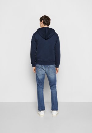 Mężczyzna w granatowej bluzie z kapturem, niebieskich jeansach i białych sportowych butach stoi twarzą do jednolitej jasnoszarej ściany, z rękami swobodnie opadającymi wzdłuż ciała.