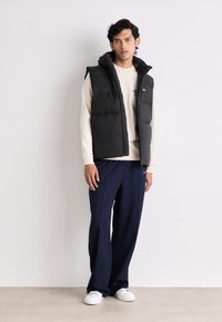 Black padded vest with a hood, featuring a zip closure. Worn over a cream sweater and paired with navy trousers and white sneakers.