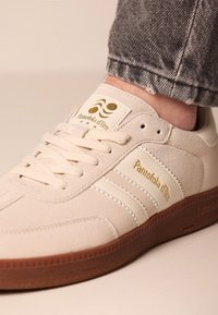 Baskets en suède beige avec des rayures blanches, semelle en caoutchouc marron et détail logo doré sur la languette et sur le côté, avec un système de laçage à l'avant et un matériau texturé.