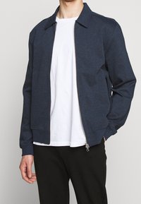 Veste zippée bleu marine en tissu texturé avec col, poches latérales et silhouette épurée. Portée sur un t-shirt blanc.