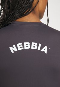 Harmaa urheilu paita, jossa on leveä, kaareva, valkoinen "NEBBIA" -logo selkäpuolella, sileää, teksturoitua kangasta ja minimaalista ompelua.