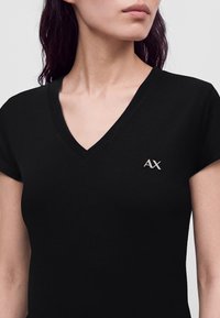 Femme portant un T-shirt noir à col en V avec un petit logo argenté AX sur le côté gauche de la poitrine sur un fond uni.