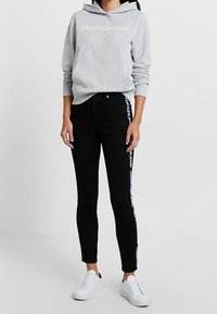 Grå huvtröja med "CALVIN KLEIN JEANS" i vit text, kombinerad med svarta skinny jeans med logodetalj på sidan och vita sneakers.
