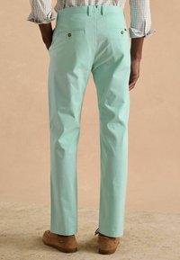 Világoszöld nadrág sima pamut felülettel, két hátsó zsebbel és barna gombdíszítéssel, barna lábbelivel párosítva.