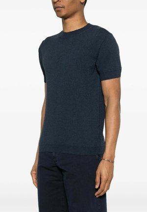 Uomo che indossa un maglione a maniche corte blu scuro e pantaloni scuri, in piedi con le braccia rilassate lungo i fianchi.