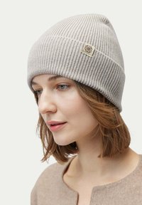 Ljusgrå ribbad mössa med vikbar bräm och en liten fyrkantig guldsymbol. Mjuk textur, något slouchig form, lämplig för kallt väder.