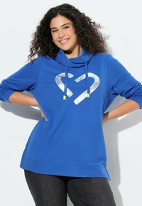 Modrá mikina s kapucí s límcem, dlouhými rukávy a dvěma velkými stříbrnými srdcovými tvary na přední straně. Má zelené akcenty na šňůrkách.