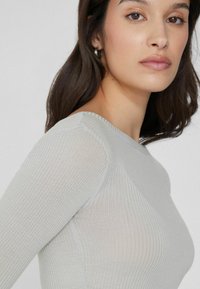 Haut en maille côtelée gris clair avec manches longues et col rond, présentant une texture lisse et une silhouette ajustée.