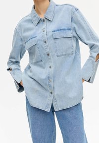 Camicia di jeans azzurro chiaro con colletto, chiusura frontale con bottoni, due tasche sul petto e maniche ampie con dettagli a taglio.