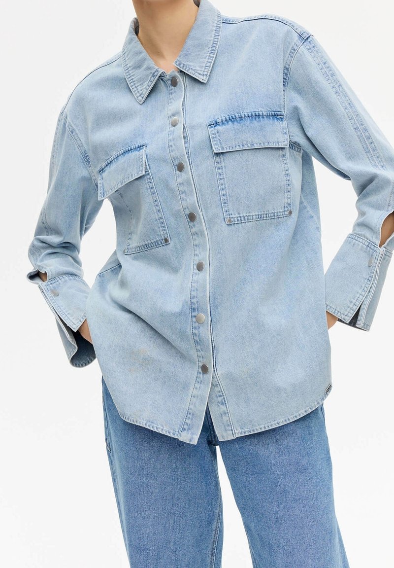 Camicia di jeans azzurro chiaro con colletto, chiusura frontale con bottoni, due tasche sul petto e maniche ampie con dettagli a taglio.