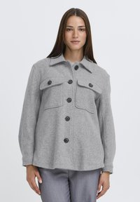 Šedá košeľa-jacket z vlnenej zmesi s golierom, dvoma náprsními vreckami a čiernymi gombíkmi. Má dlhé rukávy a voľný strih.