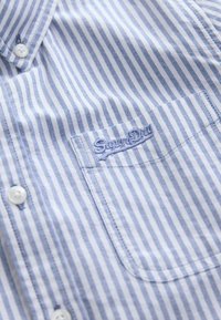 Chemise à rayures verticales bleues et blanches avec des boutons blancs et une poche avant brodée du logo "Superdry" en fil bleu.