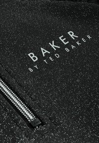 Juodas užtrauktukas striukė su tekstūruotu, spindinčiu paviršiumi. Yra baltas aprašas "BAKER BY TED BAKER" ir metalinis užtrauktukas.