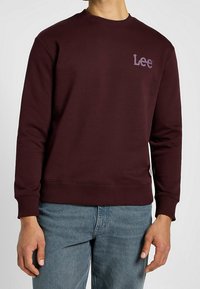 Uomo che indossa un felpa Lee di un colore borgogna scuro con polsini e orlo a coste, abbinata a jeans azzurri chiari su uno sfondo neutro.