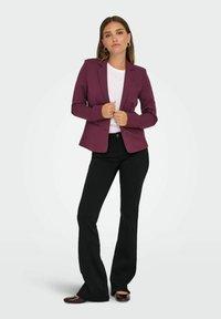 Bordeaux-farvet tætsiddende blazer med klapper, enkelt knaplukning, parret med en hvid t-shirt og sorte flared bukser, båret med mørke sko.