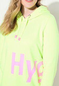 Neon groene hoodie met een voorzijdeprint in het roze met de tekst "Hy" en een trekkoord met roze accenten. Zachte, gladde textuur.