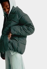 Person med en mörkgrön puffjacka över en ljusgrå hoodie och joggingsbyxor, som drar upp eller ner dragkedjan på jackfickan.