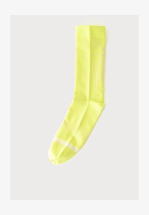 Une chaussette jaune unique avec une bande blanche près de l'orteil, posée à plat sur un fond gris clair.
