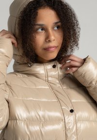 Beige pufferjack is voorzien van een glanzende, gequiltte textuur, een hoge kraag en een capuchon, beveiligd met een drukknoopsluiting en zwarte accenten.