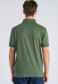 Man draagt een groene poloshirt met korte mouwen en verticale strepen, staand met de rug naar de camera, gecombineerd met een blauwe spijkerbroek tegen een effen achtergrond.