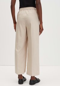 Beige wijde broek met een gestructureerde stof, elastische tailleband en twee achterzakken, gecombineerd met zwarte schoenen.