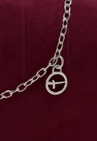 Chaîne en argent avec des maillons ovales et un pendentif circulaire avec une barre horizontale, sur un fond de tissu rouge foncé texturé.