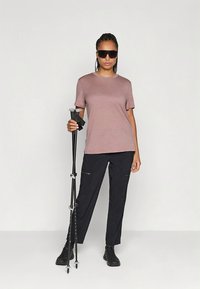 Lys lilla kortermet t-skjorte, mørke cargobukser, svarte tursko og solbriller. Holder en fjellstøvel.