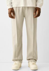 Světle béžové tepláky z měkké látky, s uvolněným střihem, jemnými záhyby a v kombinaci s krémově bílými teniskami.