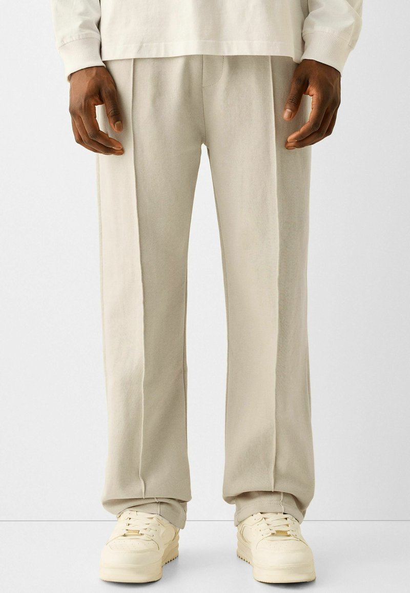 Světle béžové tepláky z měkké látky, s uvolněným střihem, jemnými záhyby a v kombinaci s krémově bílými teniskami.