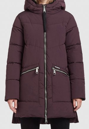 Femme portant un long manteau matelassé violet avec une fermeture éclair frontale, deux poches diagonales zippées et une capuche à col haut.