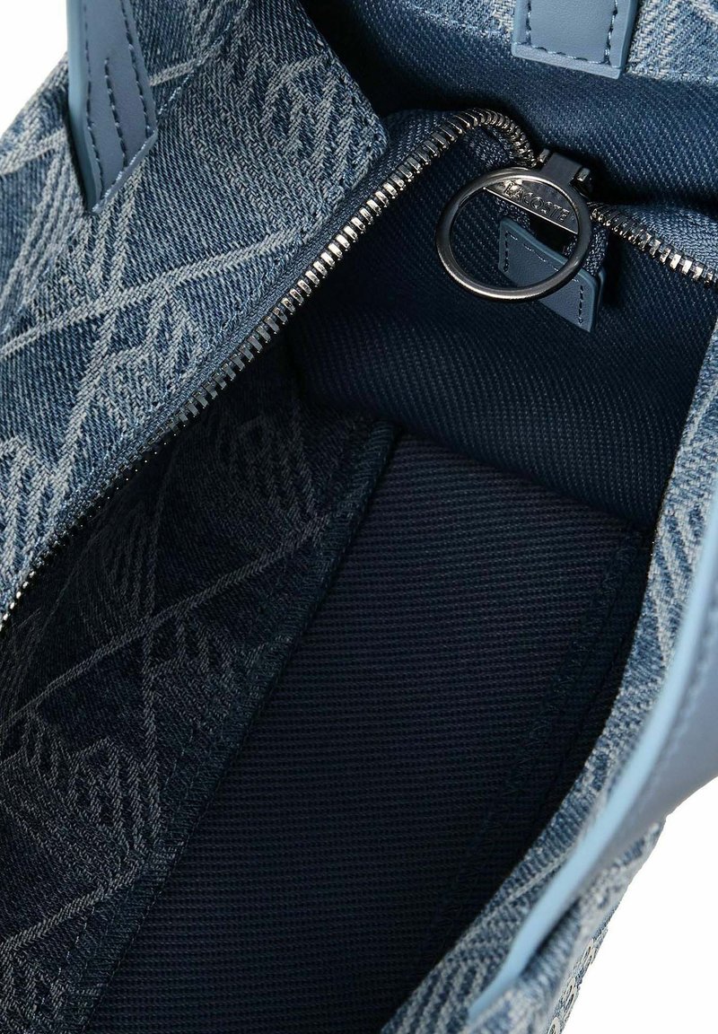 Interno di borsa con texture jeans, motivo geometrico, fodera in tessuto blu e anello argentato per la cerniera inciso con "Lacoste".