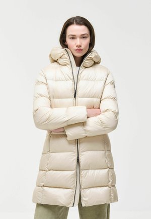Frau trägt eine cremefarbene Steppjacke mit Kapuze und steht mit verschränkten Armen vor einem einfarbigen hellen Hintergrund.