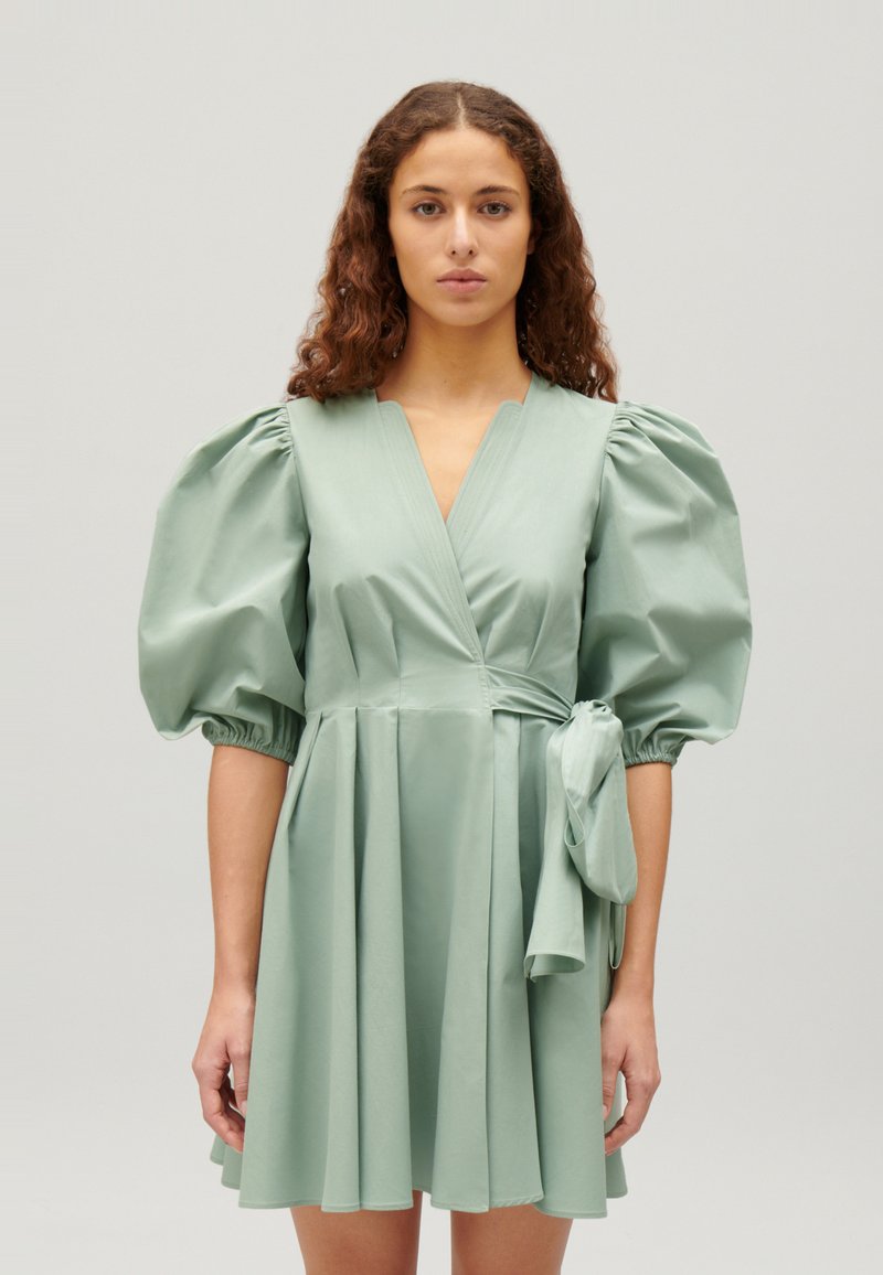 Rochie verde cu manșete voluminoase, decolteu în V și talie adunată. Fabricată dintr-un material neted, cu fustă fluidă și accent legat la talie.