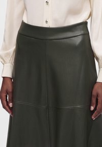 Jupe midi en cuir vert foncé avec des détails de coutures associée à un chemisier blanc avec manches bouffantes et boutons décoratifs.