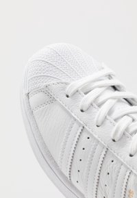 Vita lädersneaker med en texturerad tå, slät kropp och vita snörningar. Accenter inkluderar tre horisontella ränder och perforerade detaljer.