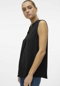 Top noir sans manches en tissu texturé avec un décolleté en V, associé à un collier en perles métalliques. Le modèle porte un jean en denim bleu.