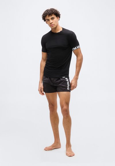 Jeune homme debout pieds nus, vêtu d'un t-shirt noir ajusté et d'un short noir avec du texte blanc sur la manche et la jambe, sur un fond clair uni.