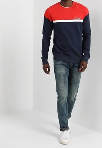 Sudadera azul marino con mangas raglán rojas y una franja blanca en el pecho, combinada con jeans azul claro y zapatillas blancas.