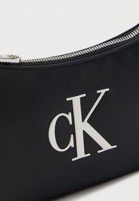 Μαύρο δερμάτινο τσαντάκι με ασημένιο φερμουάρ και μεγάλο λευκό λογότυπο “cK” στο μπροστινό μέρος, που αναδεικνύει τη λεπτομέρεια της μάρκας και την υφή.
