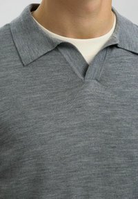 Hallide V-kaelusega kampsun halli värviga, millel on tekstuuriga kootud viimistlus. Kampsunil on kraega kaelus ja see on kerge värvusega T-särgi peale pandud.